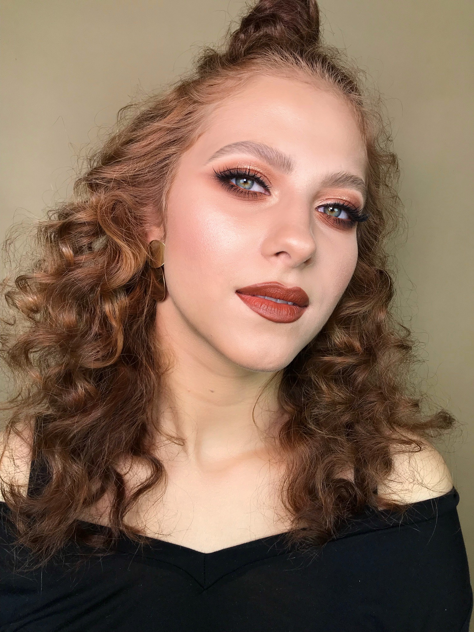 Portret kobiety z rudymi kręconymi włosami, w wyrazistym makijażu oczu w odcieniach brązu i pomadką w kolorze terakoty, prezentujący efekt końcowy usługi kosmetycznej.