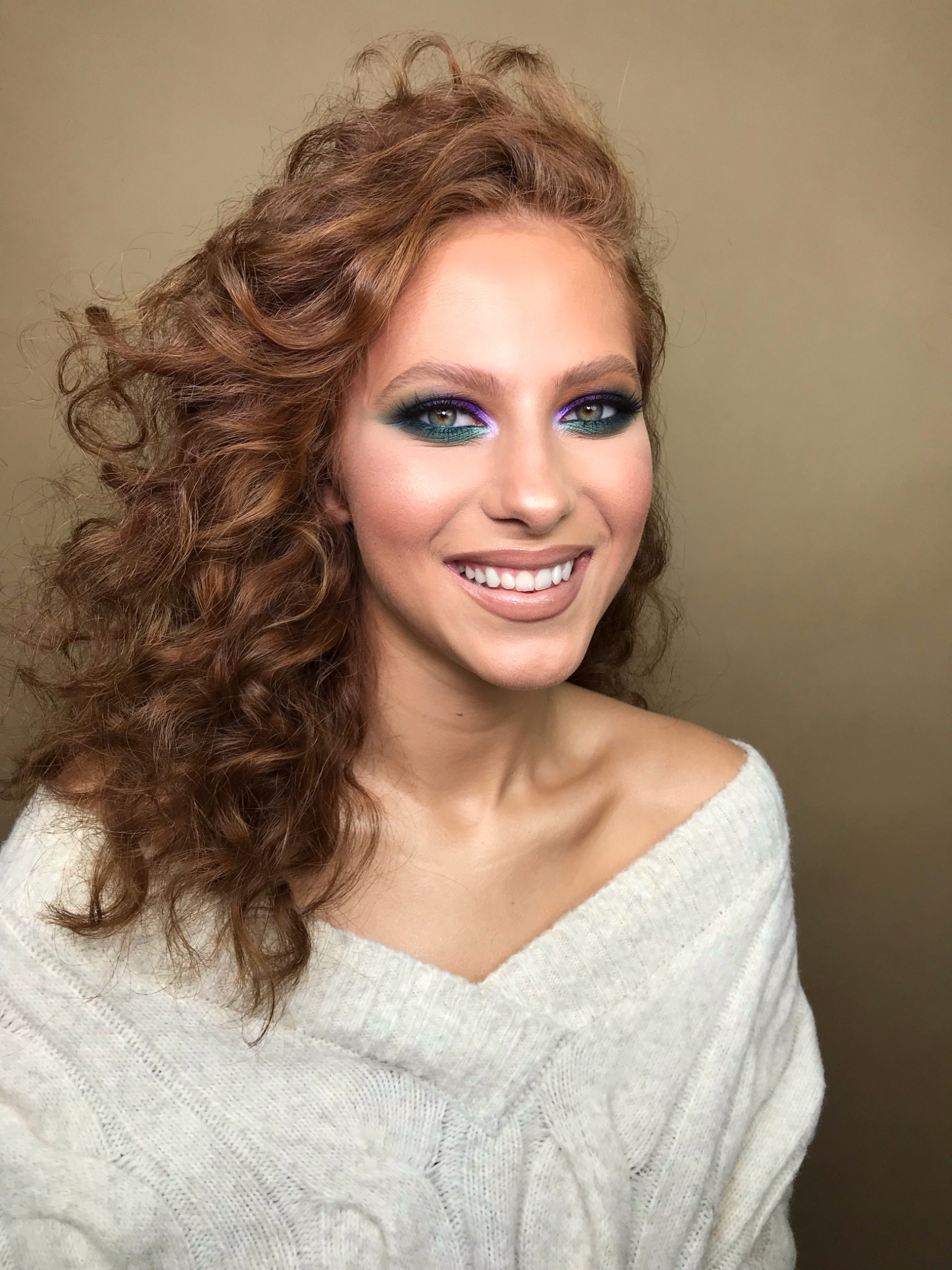 Portret uśmiechniętej kobiety z burzą rudych loków i wyrazistym makijażem oczu w odcieniach zieleni i fioletu, ubrana w biały sweter.