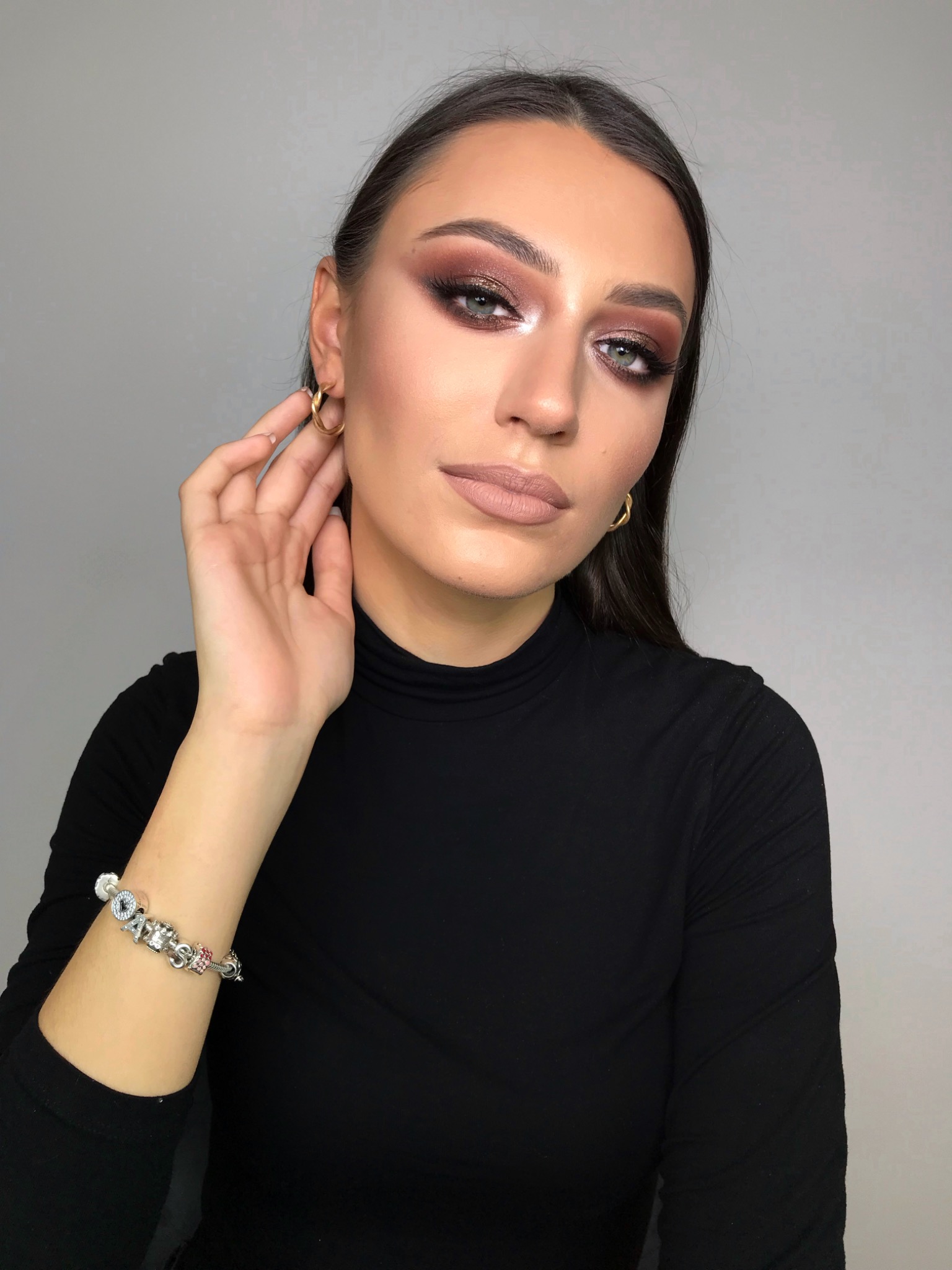 Portret kobiety z profesjonalnym makijażem oczu w odcieniach brązu i beżu, matową pomadką nude, złotymi kolczykami i bransoletką charms na ręce, pozującej na jasnoszarym tle.