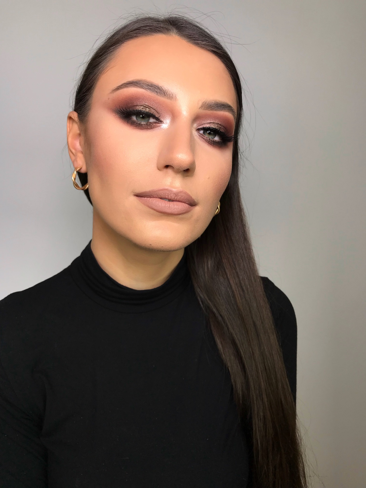 Portret kobiety z intensywnym, brązowo-złotym makijażem oczu, nude na ustach, w czarnym golfie i złotych kolczykach, z gładkimi, ciemnymi włosami opadającymi na ramię.