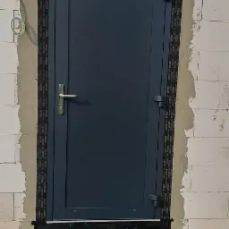 Montaż szarej, metalowej drzwi wejściowych w surowym murze z białych bloczków, zabezpieczonych taśmą uszczelniającą i częściowo obrobionych zaprawą.