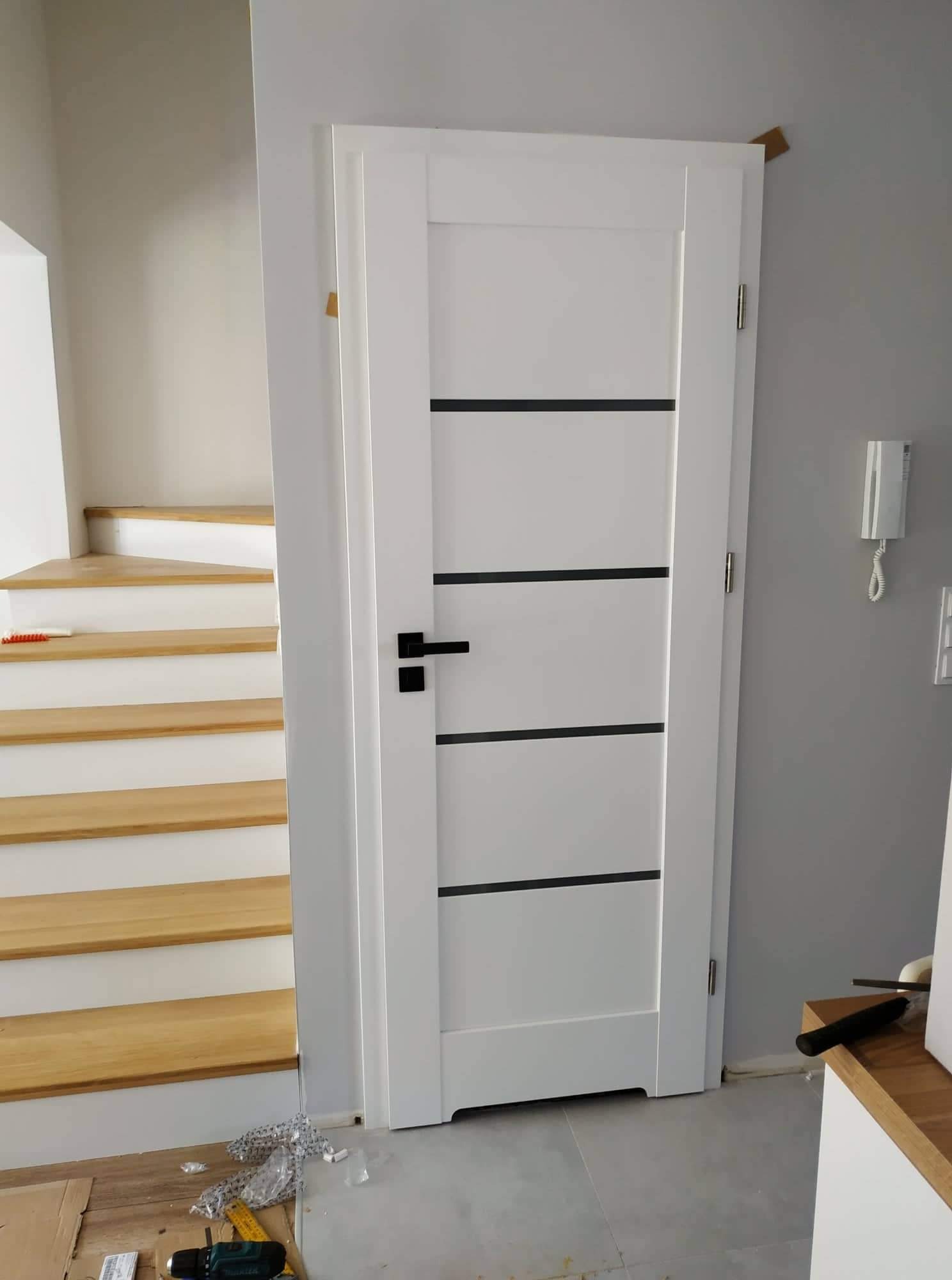 Białe drzwi wewnętrzne z czarnymi poziomymi paskami, zamontowane w nowym domu, widoczne schody z jasnego drewna i narzędzia na podłodze.