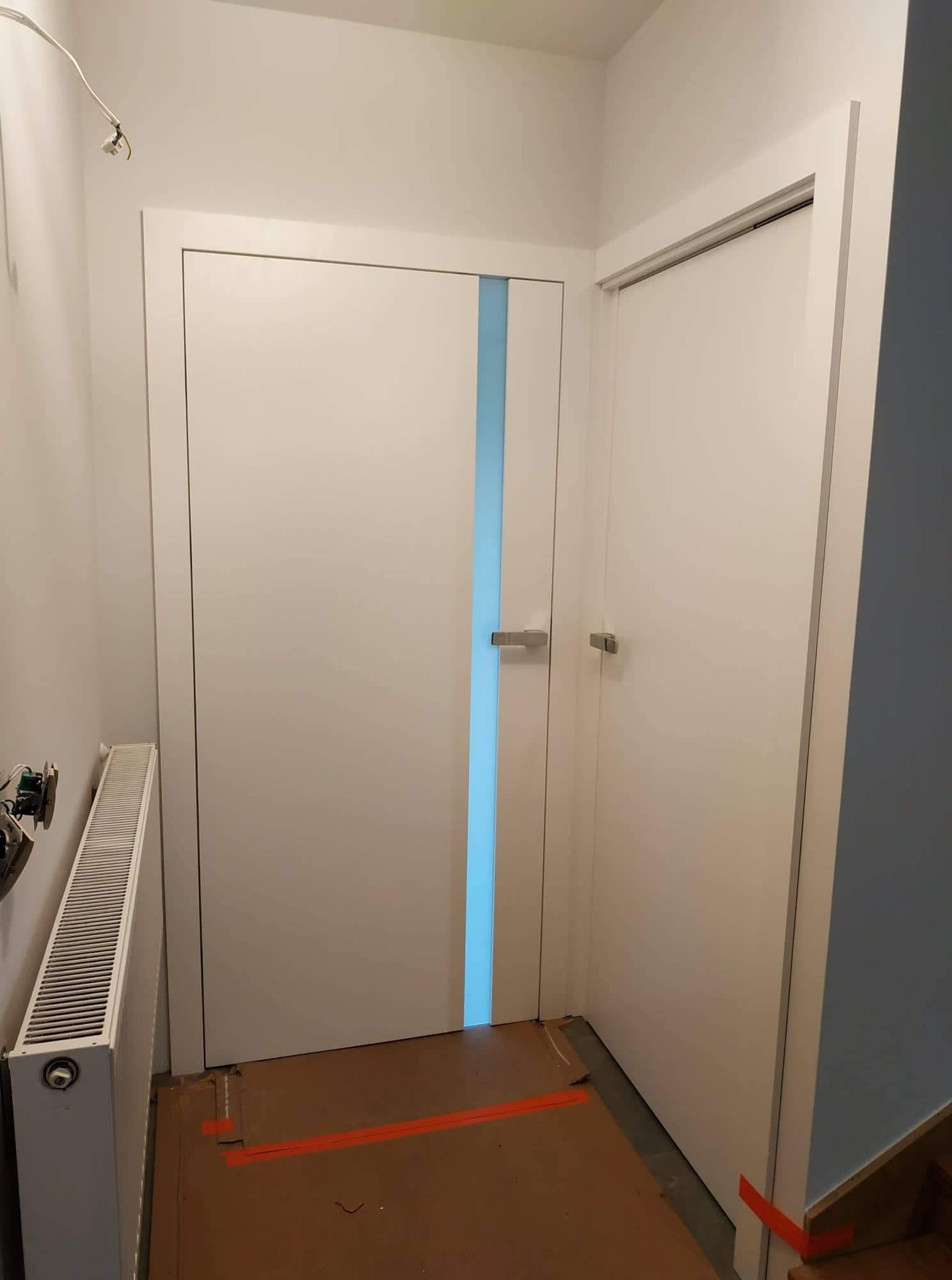 Dwa białe, nowoczesne drzwi wewnętrzne z wąskim, pionowym paskiem błękitnego szkła w jednych z nich, oraz grzejnik i instalacja elektryczna na ścianie w tle, zabezpieczona podłoga.