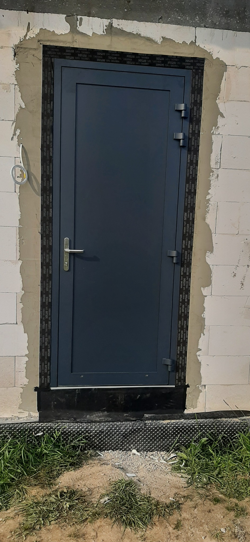 Montaż szarej, metalowej drzwi wejściowych w surowym murze z białych bloczków, zabezpieczonych taśmą uszczelniającą i częściowo obrobionych zaprawą.