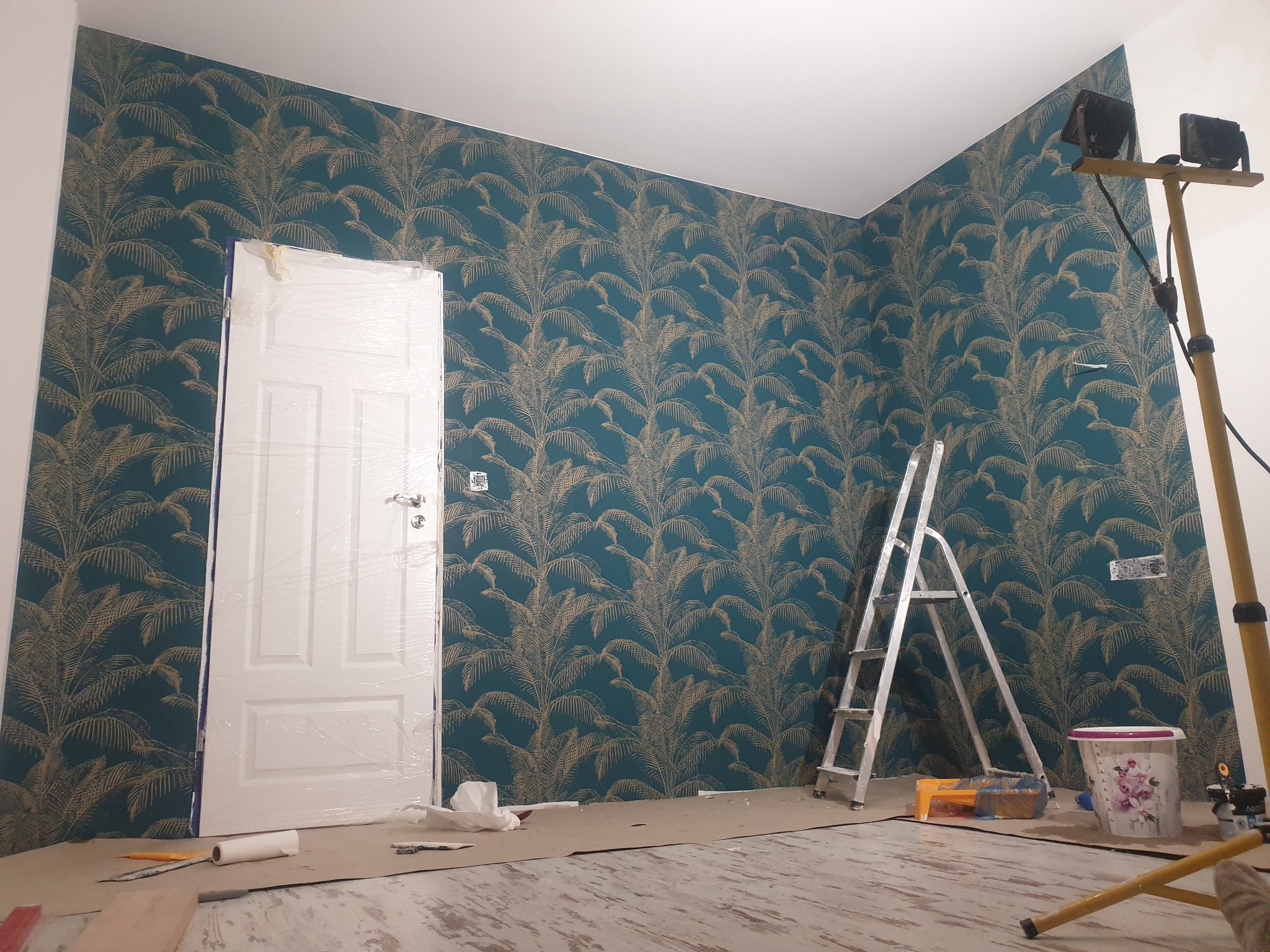 Pokój po położeniu tapety w odcieniach turkusu i złota z motywem palm, zabezpieczone folią drzwi, drabina, wiadro i lampa budowlana w trakcie prac wykończeniowych.