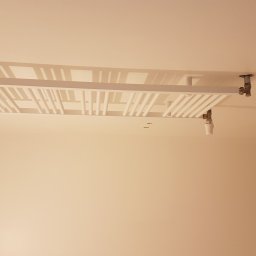 Prywatne - Nowoczesny, biały grzejnik sufitowy zamontowany w łazience w Poznaniu, widok z dołu, na tle świeżo pomalowanych ścian i surowego betonu na rogu ściany.