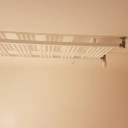 Nowoczesny, biały grzejnik sufitowy zamontowany w łazience w Poznaniu, widok z dołu, na tle świeżo pomalowanych ścian i surowego betonu na rogu ściany.