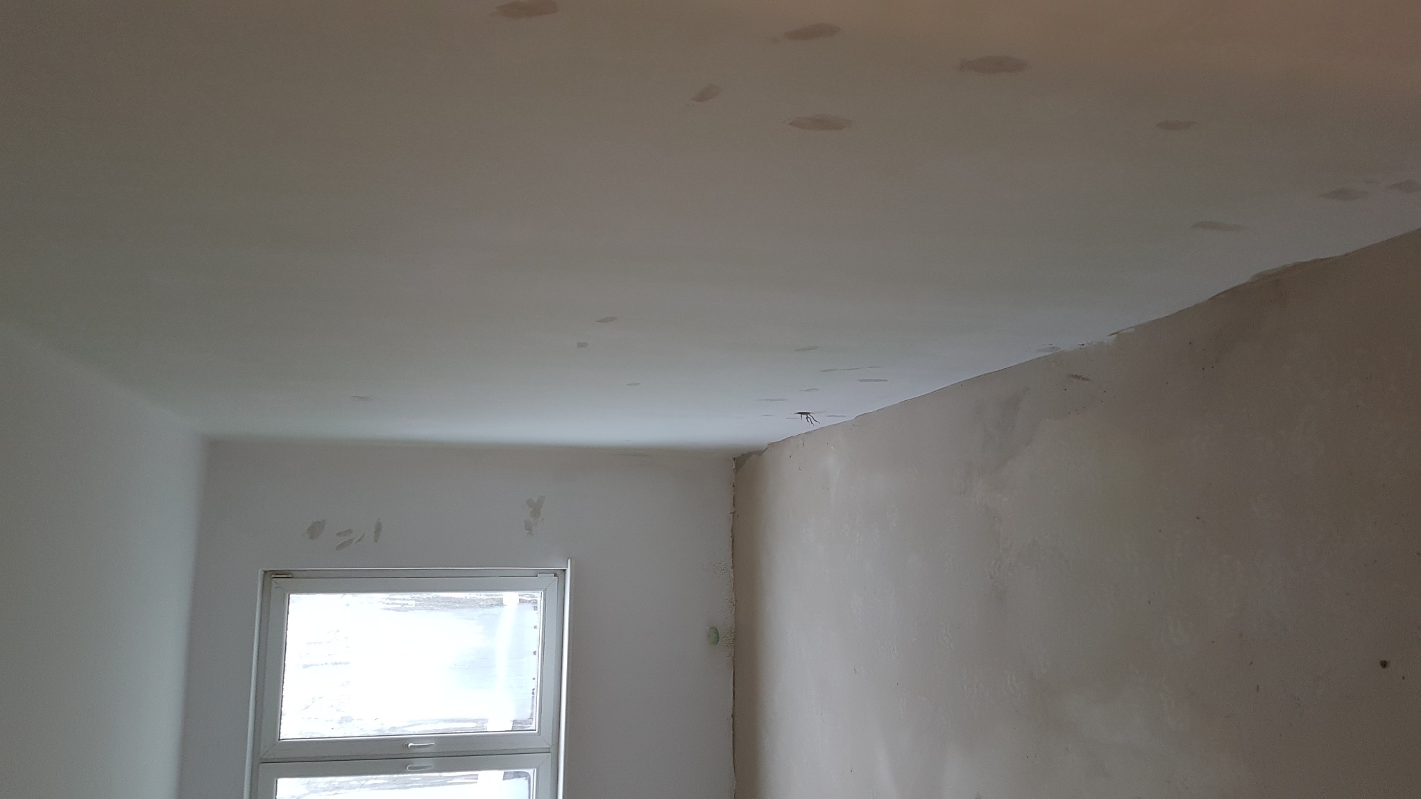 Pokój w trakcie szpachlowania: widoczne ślady po łatach na suficie i ścianach, okno z białymi ramami w tle.