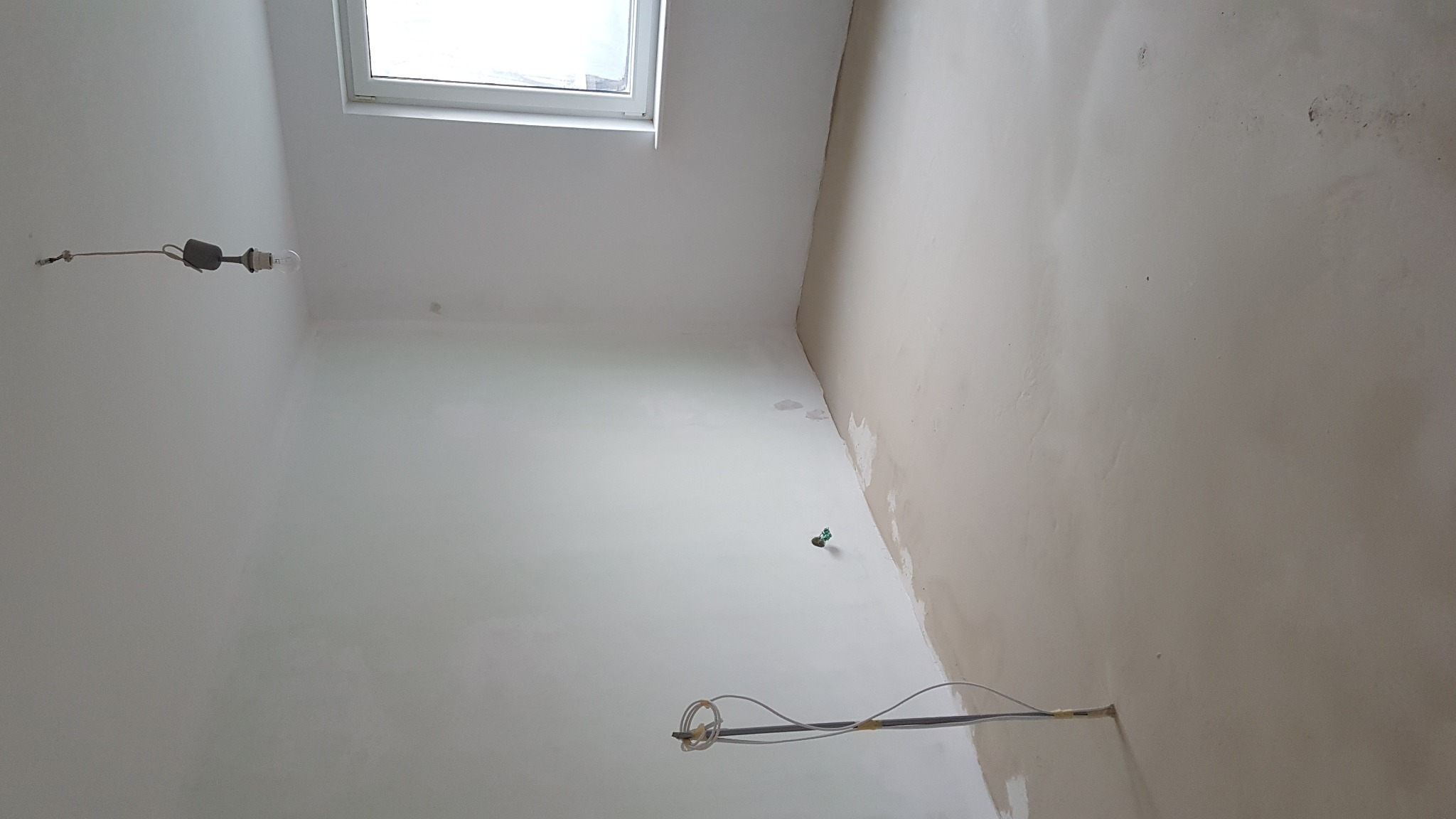 Pokój w trakcie remontu: ściany częściowo wyszpachlowane, widoczne okno, wisząca żarówka i przewody elektryczne wystające ze ściany.