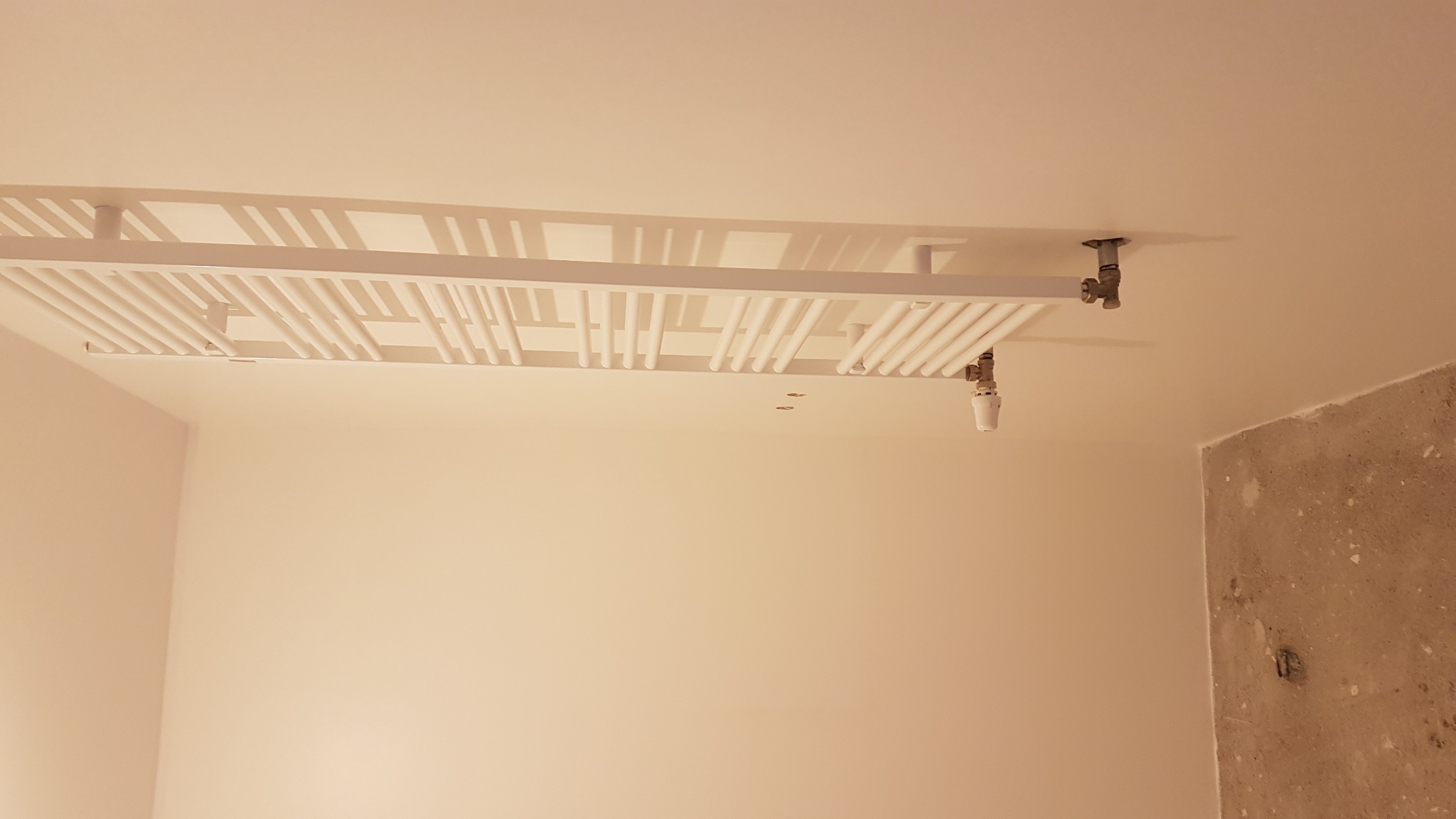 Nowoczesny, biały grzejnik sufitowy zamontowany w łazience w Poznaniu, widok z dołu, na tle świeżo pomalowanych ścian i surowego betonu na rogu ściany.