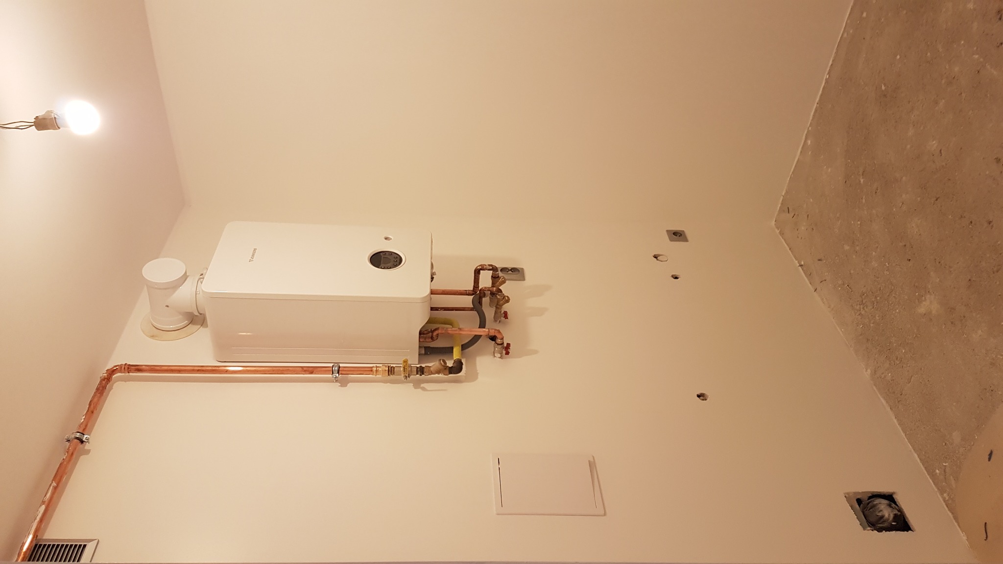 Biały piec gazowy zamontowany na ścianie z miedzianymi rurami, widoczne ślady po gniazdkach i surowy fragment ściany w rogu.