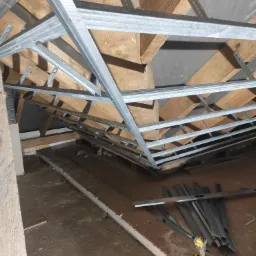 Stelaż z profili metalowych zamontowany do drewnianej konstrukcji poddasza, widoczny poziomica i wkrętarka akumulatorowa. Przygotowanie do ocieplenia.