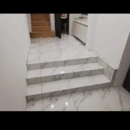 Wyłożone marmuropodobnymi płytkami schody z trzema stopniami, prowadzące do drewnianych schodów, z grzejnikiem na ścianie w tle.