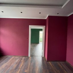 GBK - Pomalowane na bordowo ściany w pustym pokoju z drewnianą podłogą, widok na zielony pokój przez białe futryny drzwi.