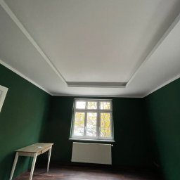 GBK - Pokój po malowaniu ścian na ciemnozielony kolor, z białym sufitem z dekoracyjnymi listwami, drewnianą podłogą, białym grzejnikiem pod oknem i małym stolikiem przy ścianie.