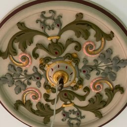 GBK - Ozdobny, okrągły element dekoracyjny sufitu z motywami roślinnymi w odcieniach zieleni, szarości, różu i złota, z widocznym przewodem do lampy w centralnej części.