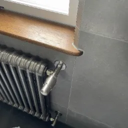 Szary grzejnik żeberkowy z chromowanymi zaworami, zamontowany pod drewnianym parapetem w łazience wyłożonej szarymi płytkami.