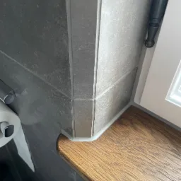 Szary, ceramiczny narożnik z płytek w łazience, połączony z drewnianym blatem okiennym i białą ramą okna, z widocznym uchwytem na papier toaletowy.