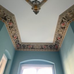 GBK - Odrestaurowany sufit w zabytkowym wnętrzu z dekoracyjnymi sztukateriami w odcieniach bieli, złota i różu, widok z dołu na błękitne ściany i okno z białymi ramami.