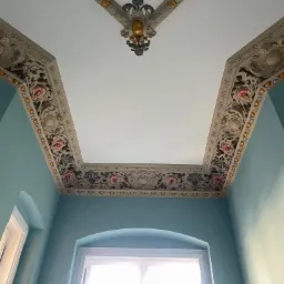 Odrestaurowany sufit w zabytkowym wnętrzu z dekoracyjnymi sztukateriami w odcieniach bieli, złota i różu, widok z dołu na błękitne ściany i okno z białymi ramami.