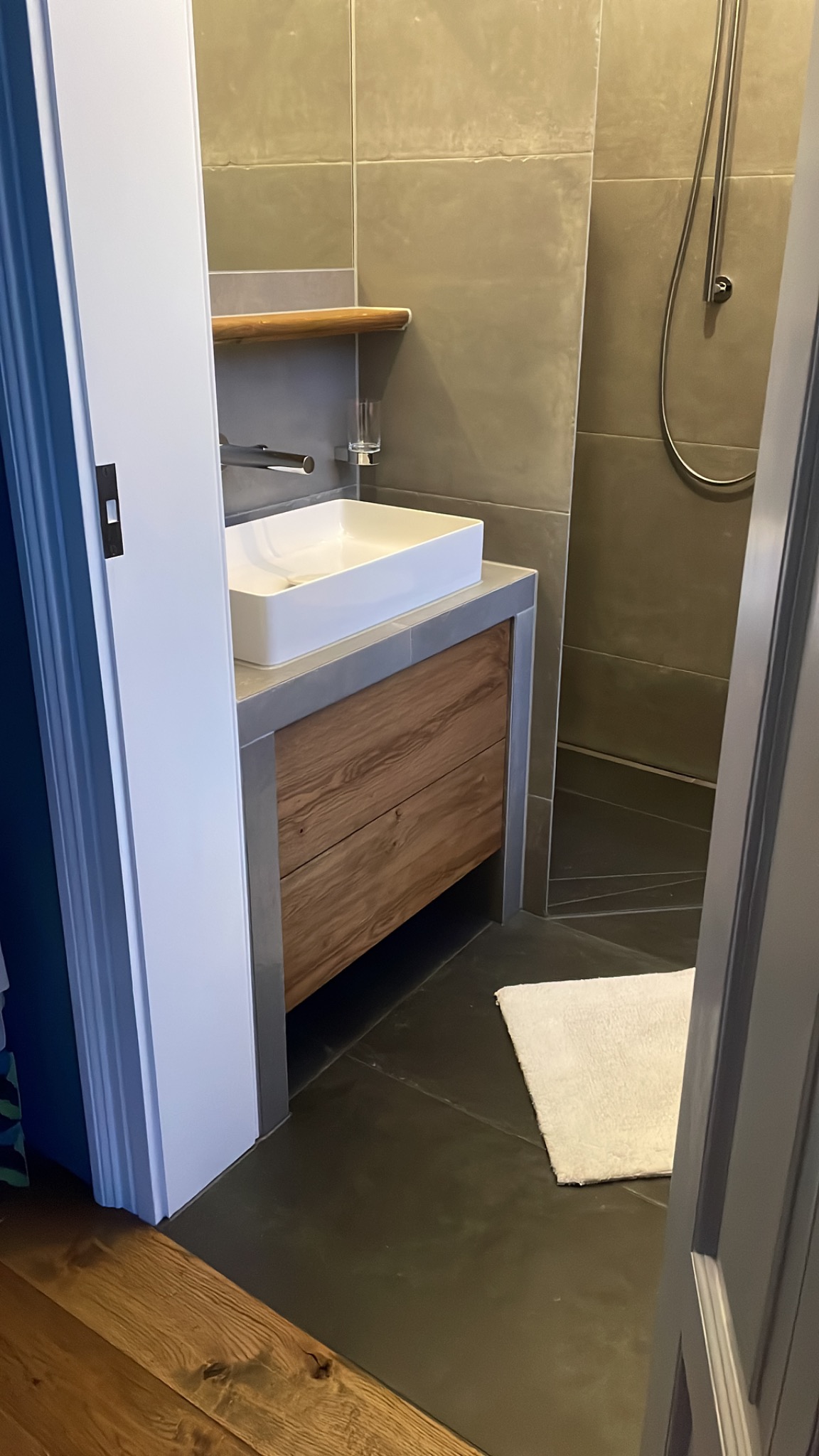 Nowoczesna łazienka z kwadratową umywalką nablatową, drewnianą szafką pod umywalką, szarymi płytkami na ścianach i podłodze oraz widoczną kabiną prysznicową z deszczownicą.