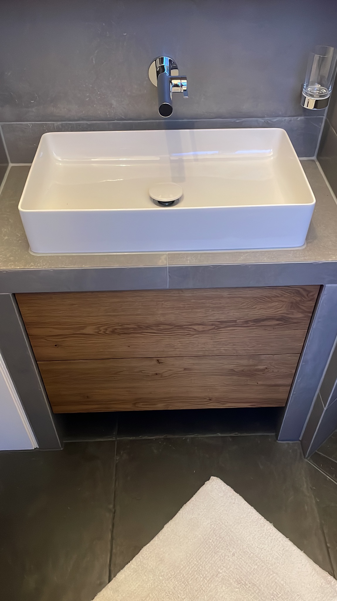 Nowoczesna łazienka z prostokątną białą umywalką nablatową, chromowaną baterią i szafką z frontem z naturalnego drewna o wyraźnym usłojeniu, na tle szarych płytek.