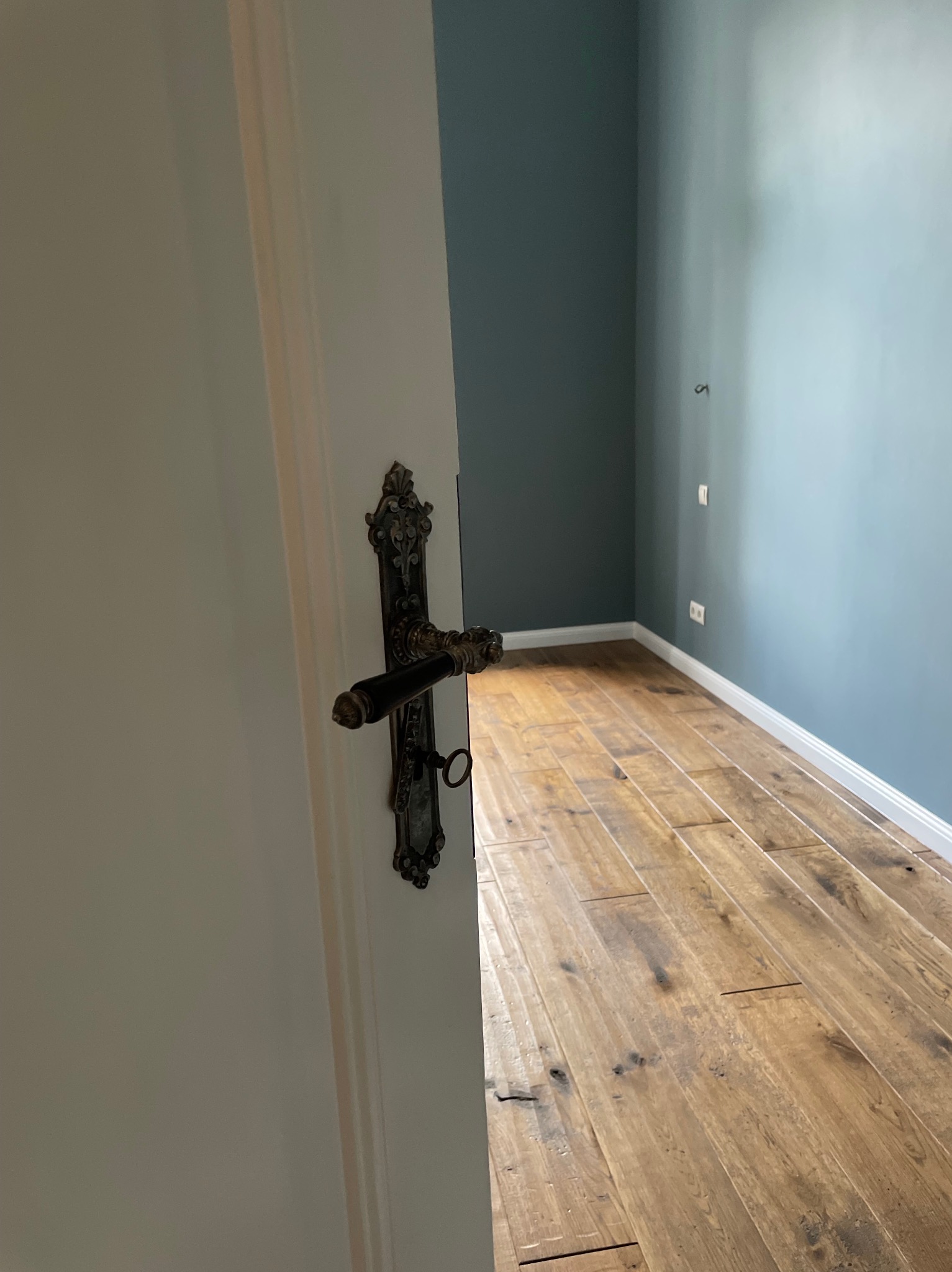 Otwarta biała drzwi z ozdobną, starą klamką, ukazująca fragment pokoju z drewnianą podłogą i pomalowanymi na niebiesko ścianami.