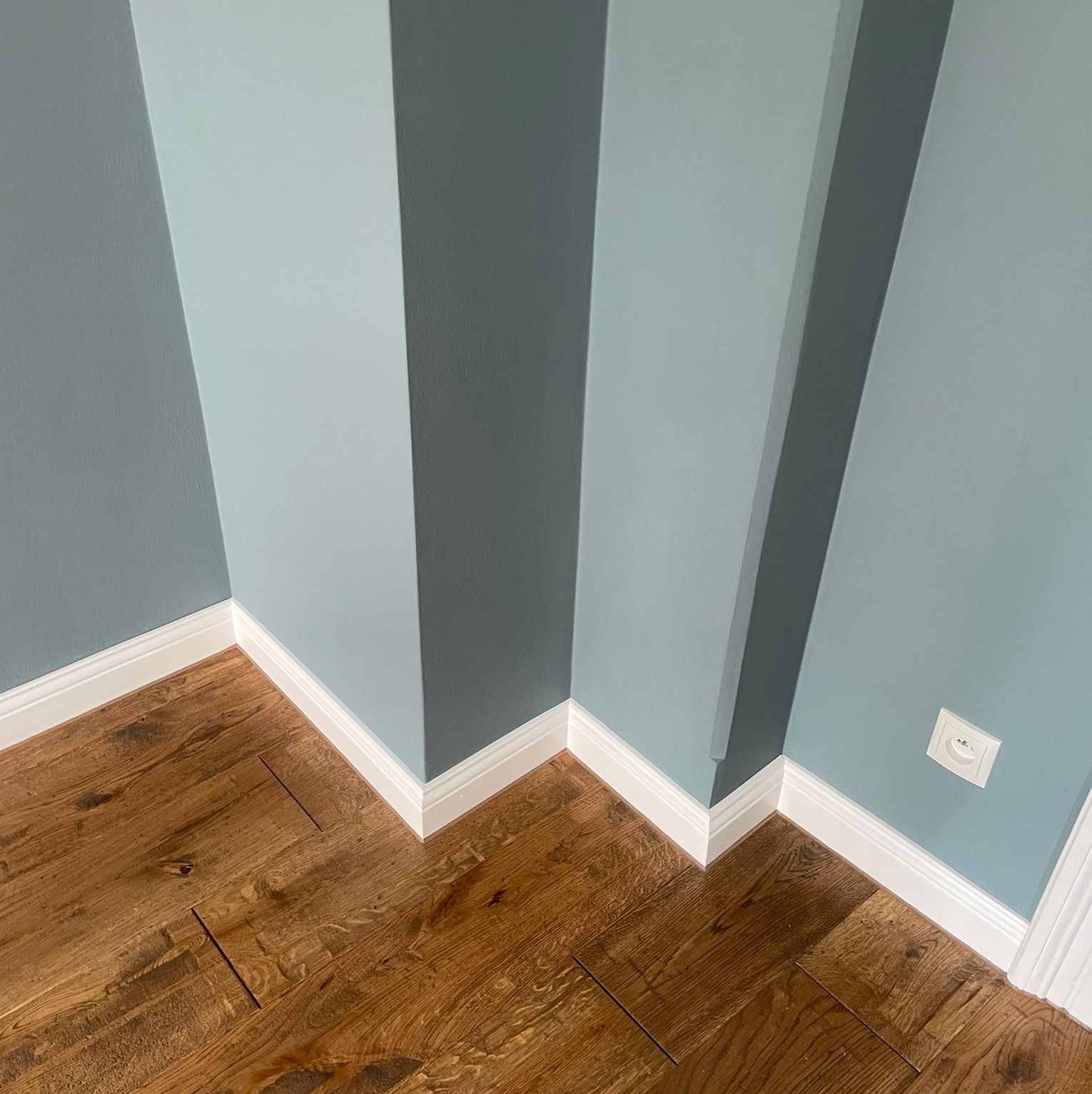Narożnik pokoju z drewnianą podłogą, błękitnymi ścianami z białymi listwami przypodłogowymi i gniazdkiem elektrycznym