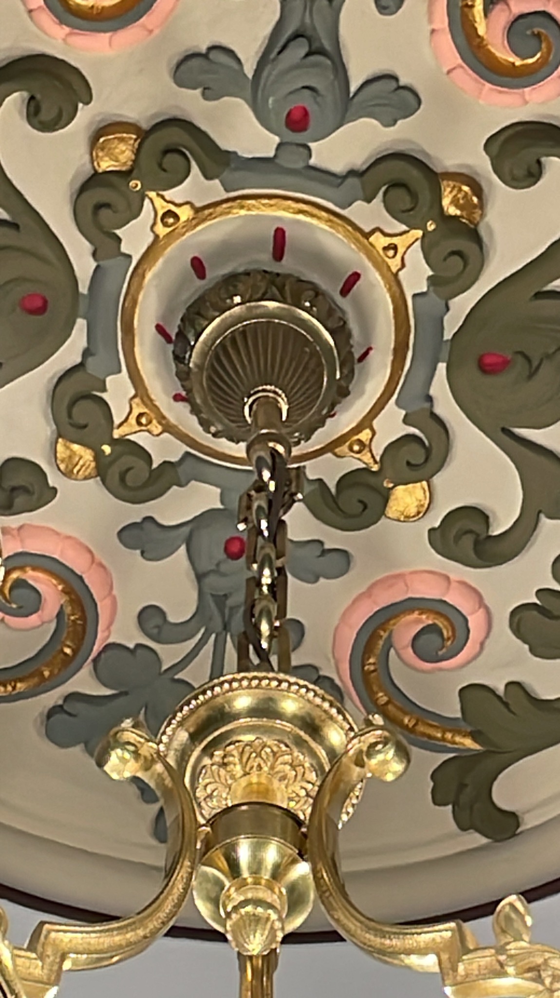 Zdobiony sufit z ornamentami w odcieniach szarości, zieleni, różu i złota, z widocznym fragmentem złotego żyrandola z łańcuchem, patrząc z dołu.