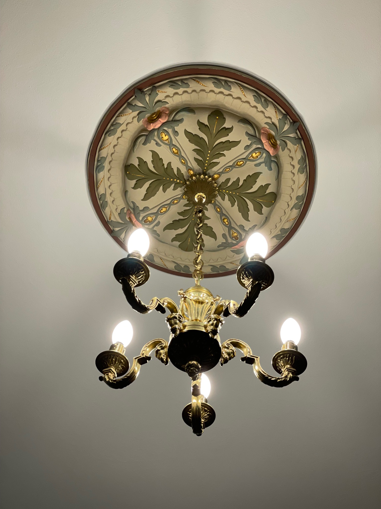 Złoty żyrandol z pięcioma ramionami i żarówkami, zawieszony pod okrągłym, ozdobnym plafonem z motywami roślinnymi i kwiatowymi na białym suficie.