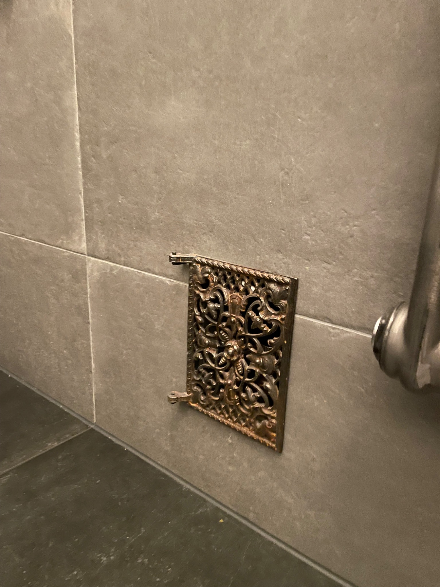 Ozdobna, metalowa kratka wentylacyjna, z ażurowym wzorem roślinnym, zamontowana na szarej, płytkowej ścianie w łazience, obok fragmentu metalowej rurki.