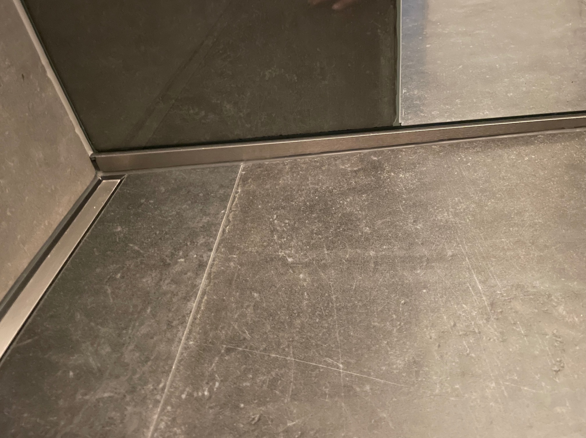 Szare płytki podłogowe z metalową listwą przy kabinie prysznicowej, widoczne delikatne zarysowania na powierzchni płytek.