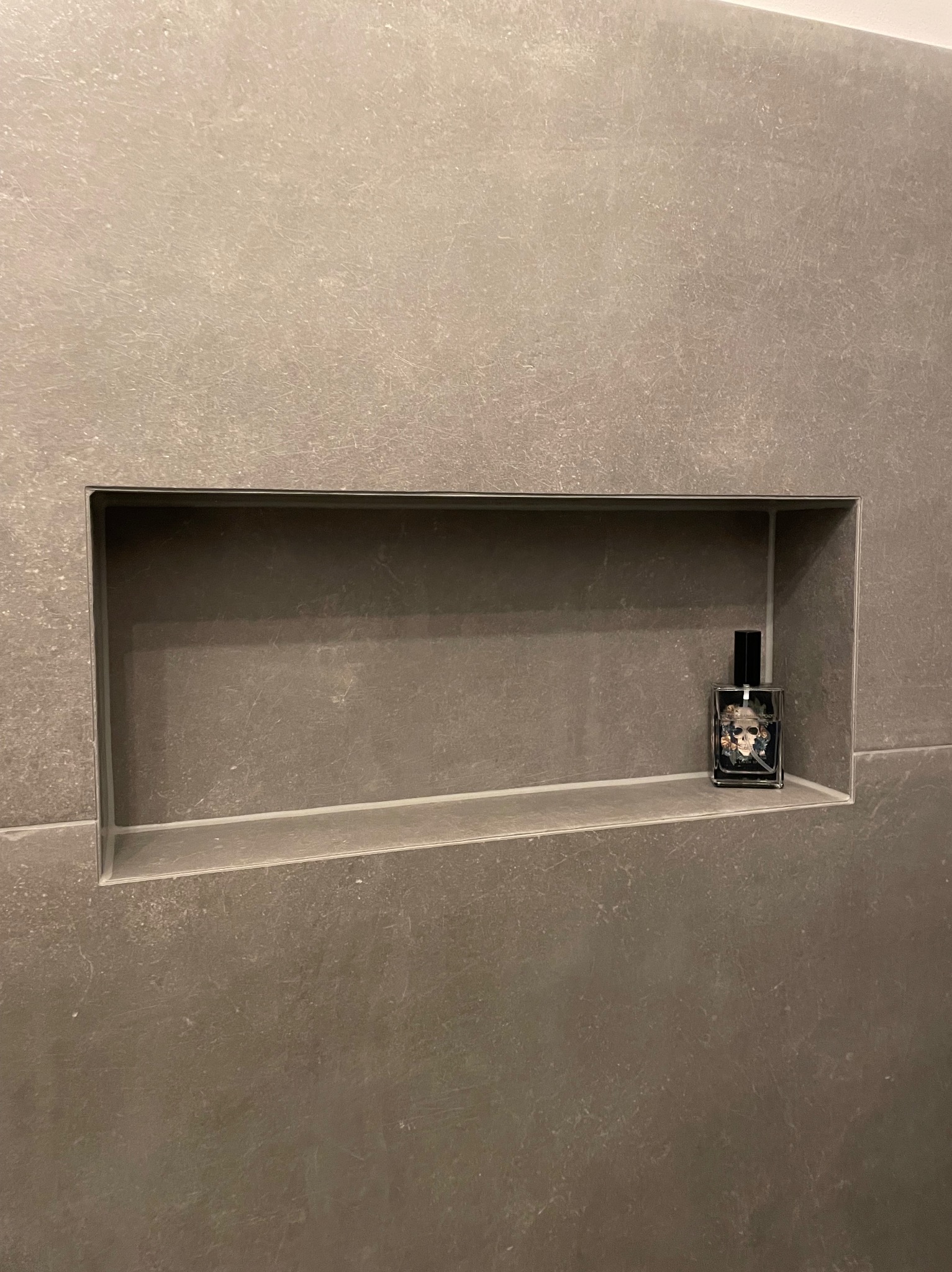 Wnęka w szarej, matowej płytce ściennej, z flakonem perfum z motywem czaszki w środku.