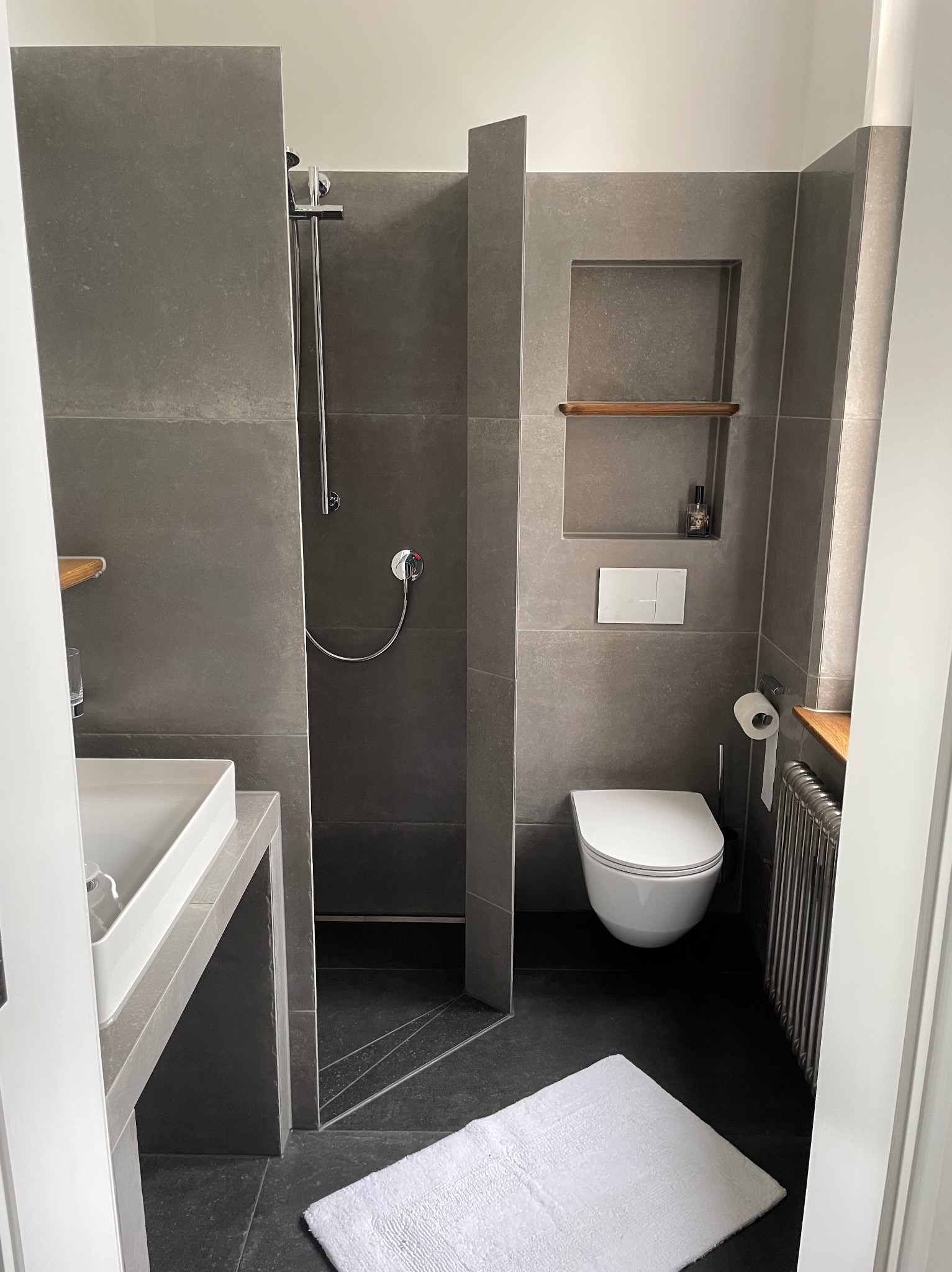 Nowoczesna łazienka z szarymi płytkami, kabiną prysznicową typu walk-in, wiszącą toaletą, grzejnikiem żeberkowym i białą matą podłogową.