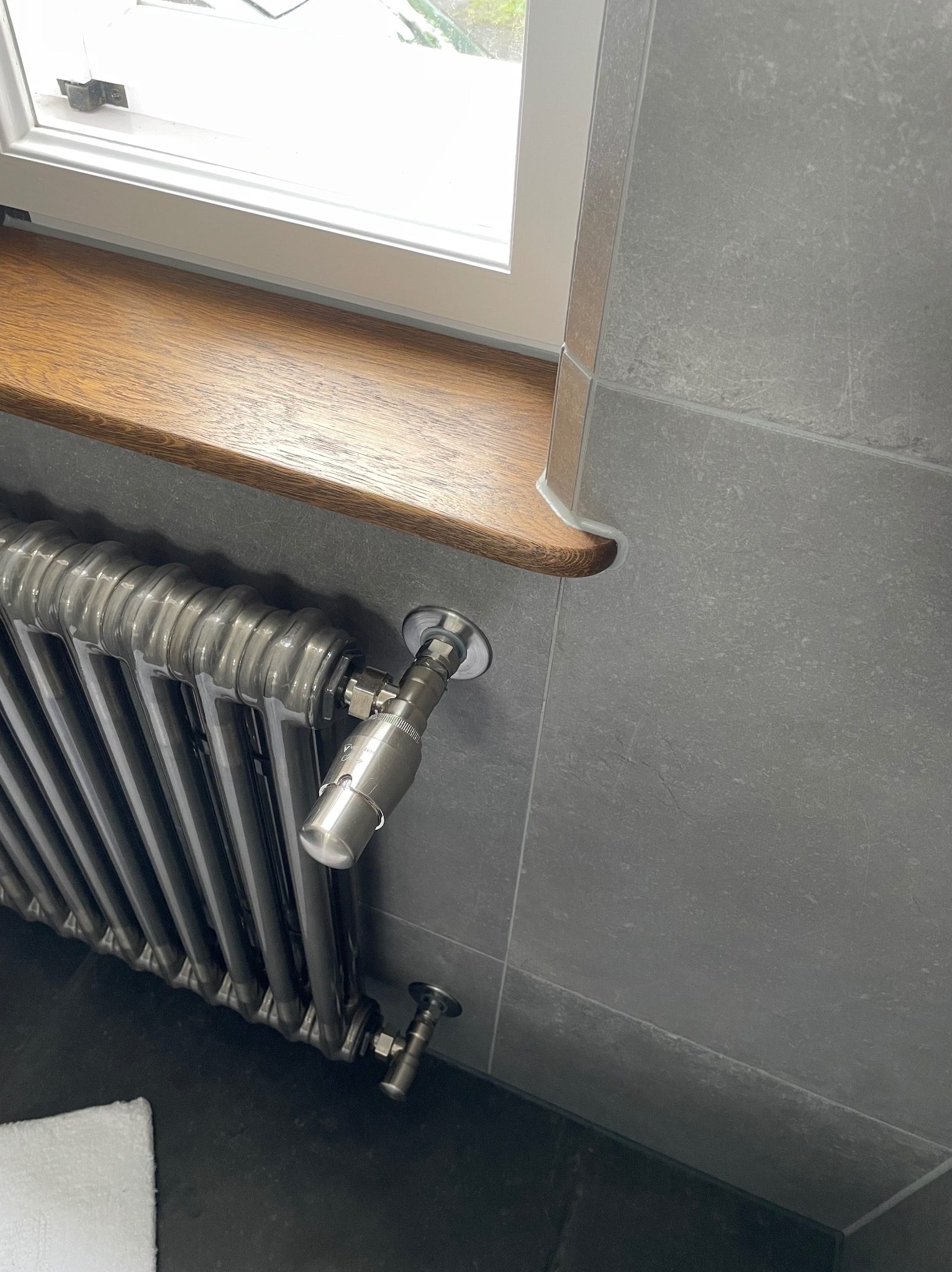 Szary grzejnik żeberkowy z chromowanymi zaworami, zamontowany pod drewnianym parapetem w łazience wyłożonej szarymi płytkami.