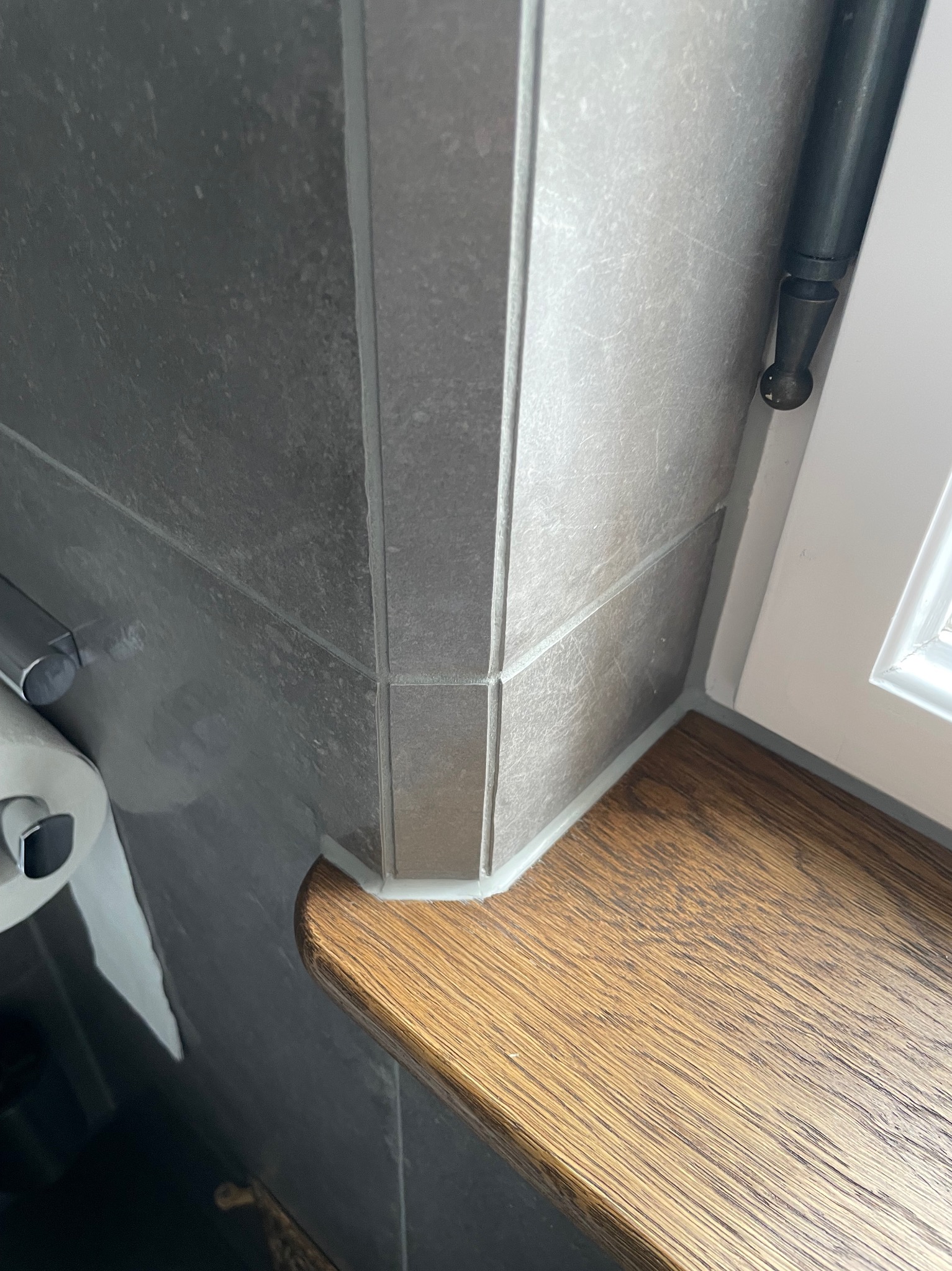 Szary, ceramiczny narożnik z płytek w łazience, połączony z drewnianym blatem okiennym i białą ramą okna, z widocznym uchwytem na papier toaletowy.