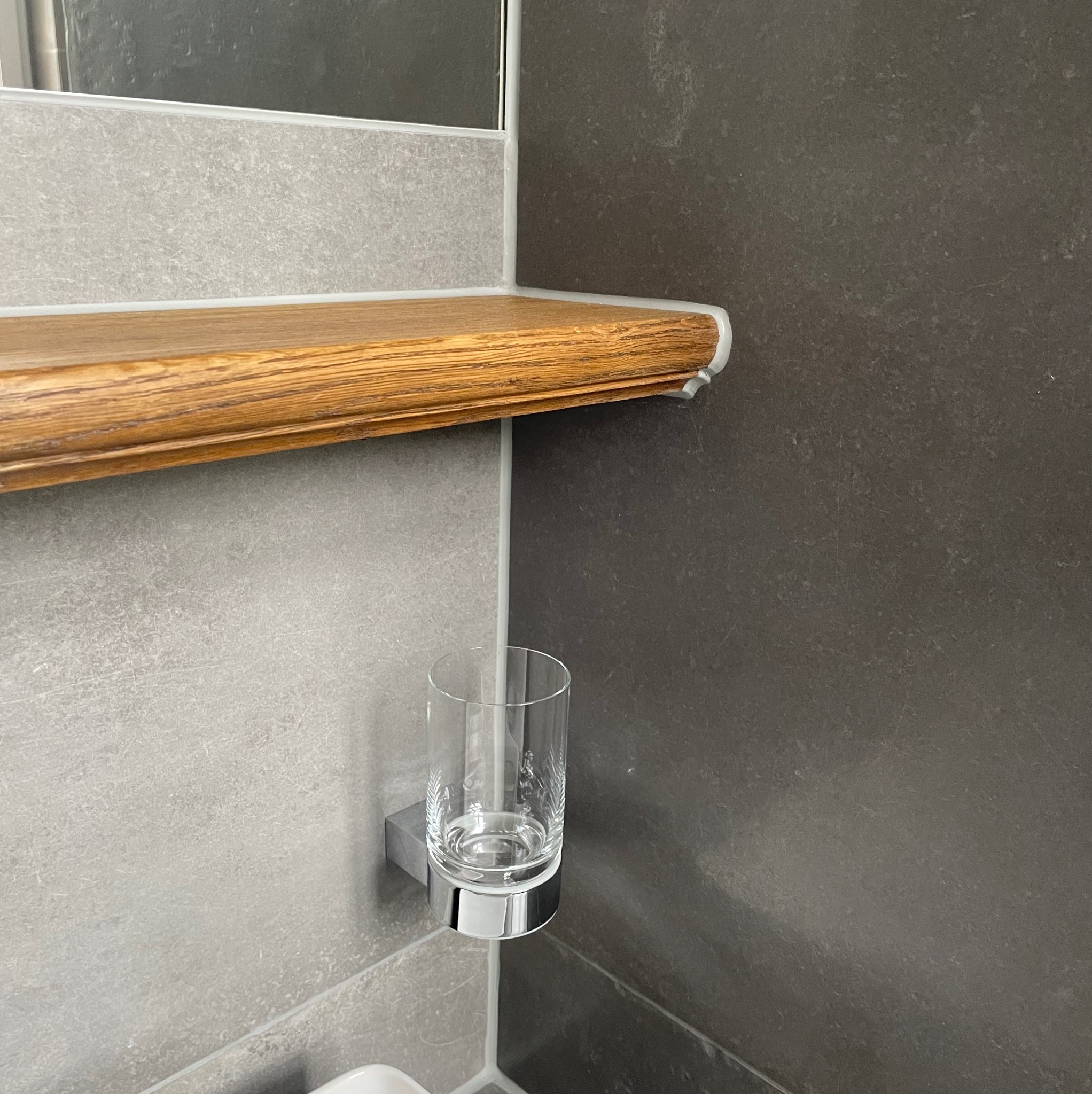 Nowoczesna łazienka: drewniana półka z ozdobnym wykończeniem, szklanka w chromowanym uchwycie na tle szarych płytek w dwóch odcieniach.