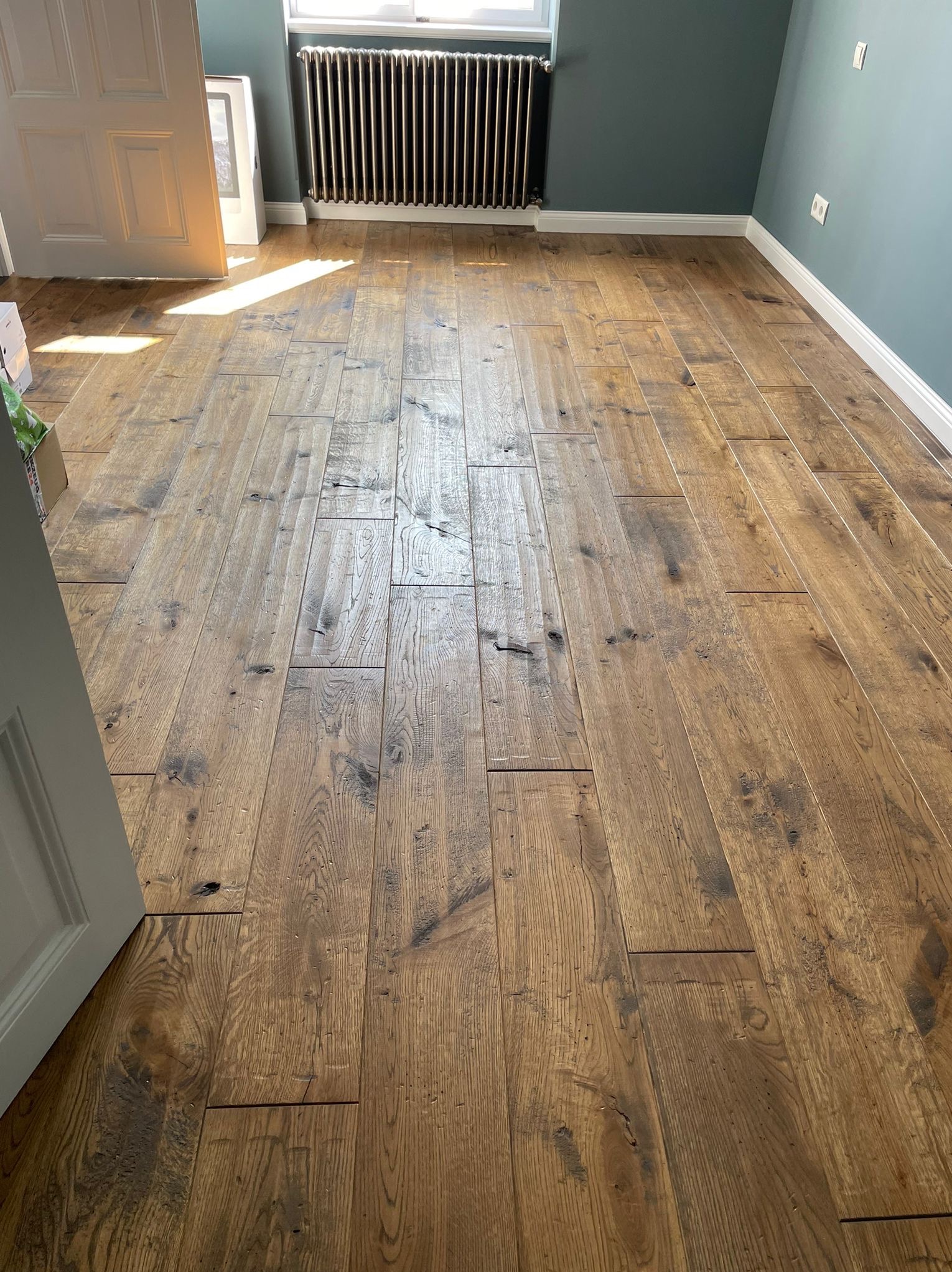 Ułożona, rustykalna podłoga z desek w pokoju z szarozielonymi ścianami, widoczny grzejnik i fragment białych drzwi.