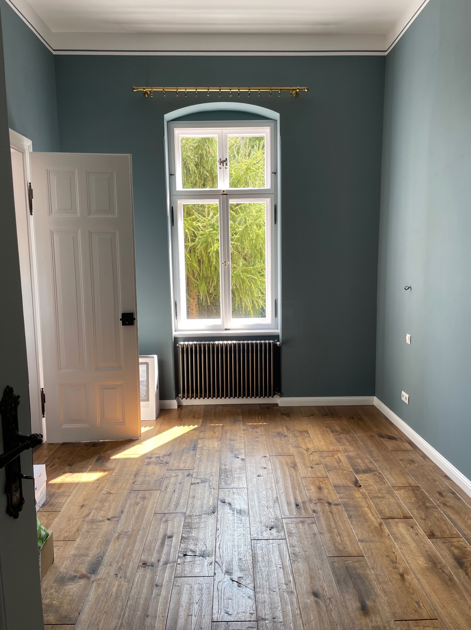 Pusty pokój po remoncie z drewnianą podłogą, pomalowanymi na niebiesko ścianami i białymi listwami przypodłogowymi. W oknie widok na zieleń. Drzwi białe, otwarte.