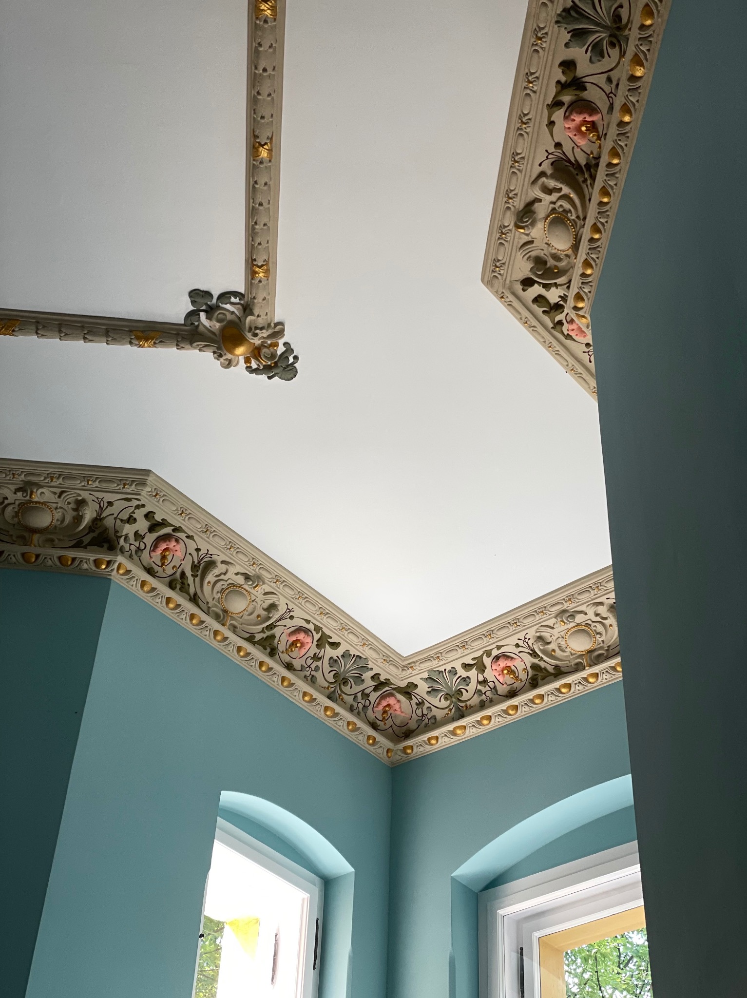 Turkusowe ściany w pokoju z białym sufitem i ozdobnymi, kremowo-złotymi sztukateriami z motywami roślinnymi i różowymi akcentami, widoczne okna z białymi ramami.