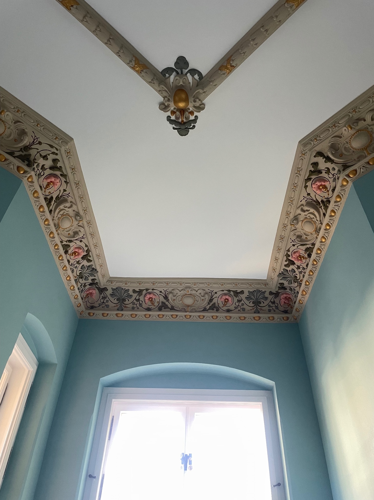 Odrestaurowany sufit w zabytkowym wnętrzu z dekoracyjnymi sztukateriami w odcieniach bieli, złota i różu, widok z dołu na błękitne ściany i okno z białymi ramami.