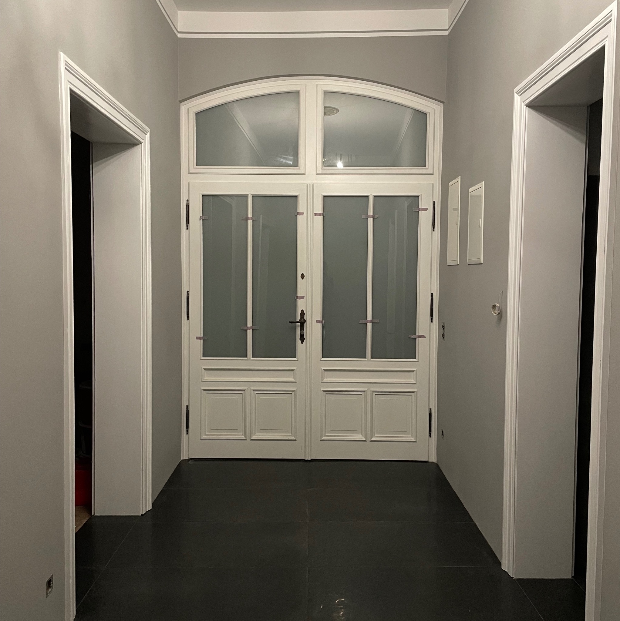 Korytarz w szarościach z czarną podłogą, białe drzwi dwuskrzydłowe z przeszkleniami zabezpieczone taśmą malarską, białe ościeżnice drzwi po bokach.