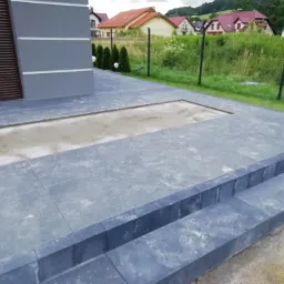 Szare płytki tarasowe ułożone na zewnątrz domu, z widocznym fragmentem przygotowanego podłoża w miejscu przyszłego trawnika lub rabaty.