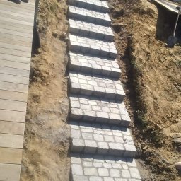 Mariusz Tymiński - Długie schody z kostki, prowadzące po stromej, piaszczystej skarpie, obok drewnianego tarasu. Widoczne buty robocze i częściowo odsłonięta ziemia.