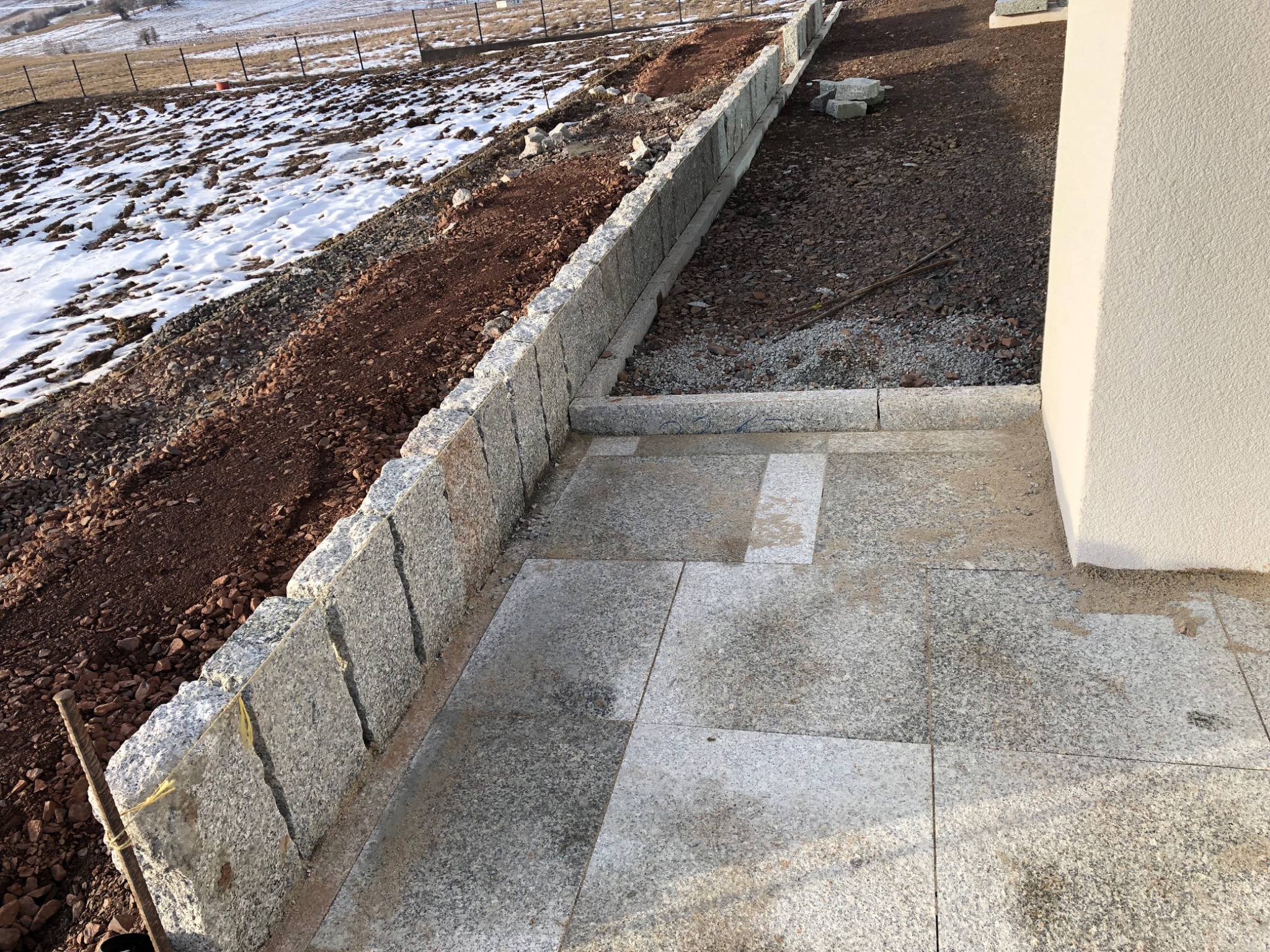 Granitowa ścieżka obok ściany budynku, z widokiem na zaśnieżone pole w tle, częściowo ukończona