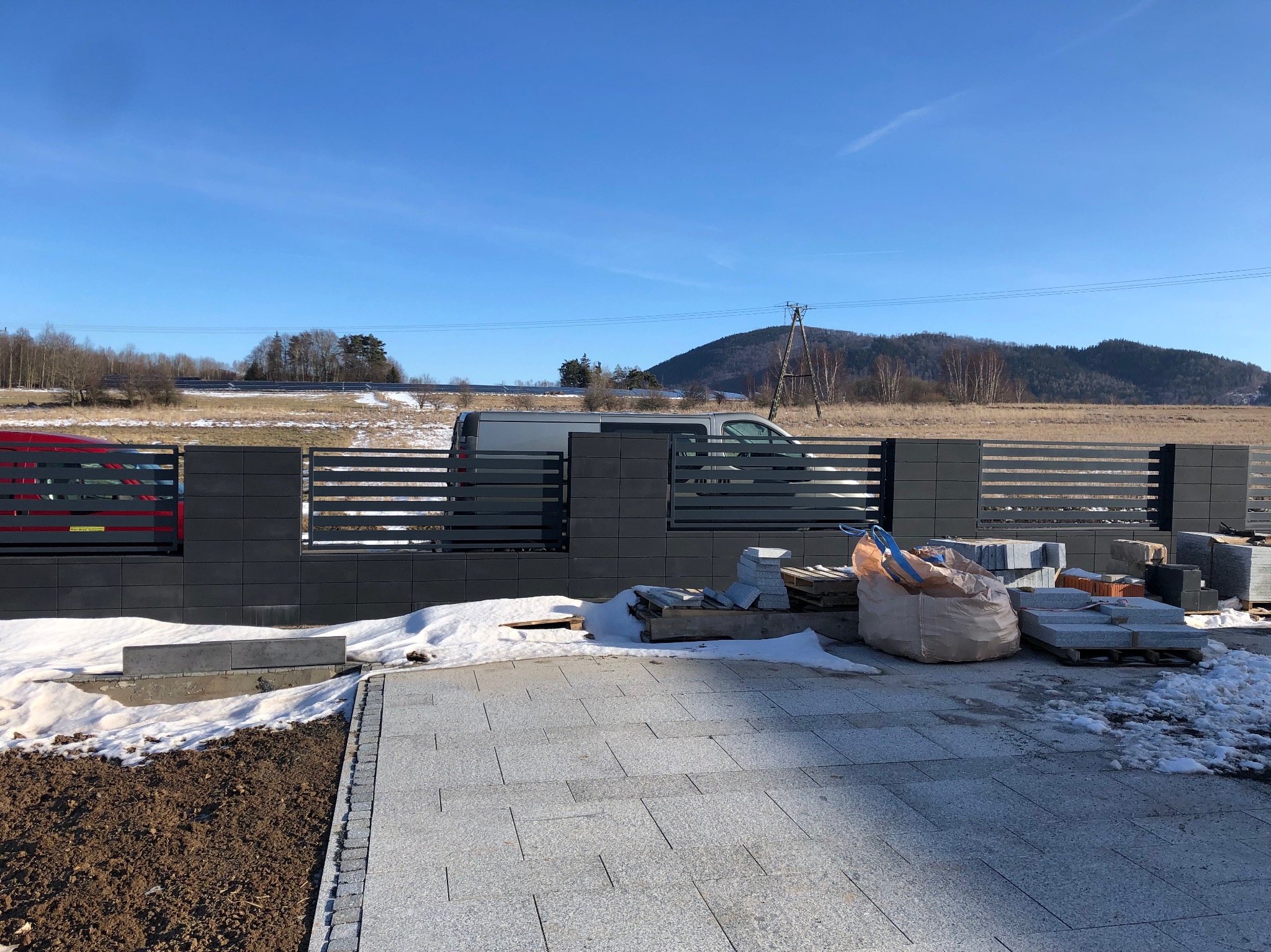 Układanie szarej kostki brukowej na podjeździe w trakcie prac, widoczny częściowo zbudowany, nowoczesny, szary płot z poziomymi przęsłami, materiały budowlane na paletach, w tle pole i góra...