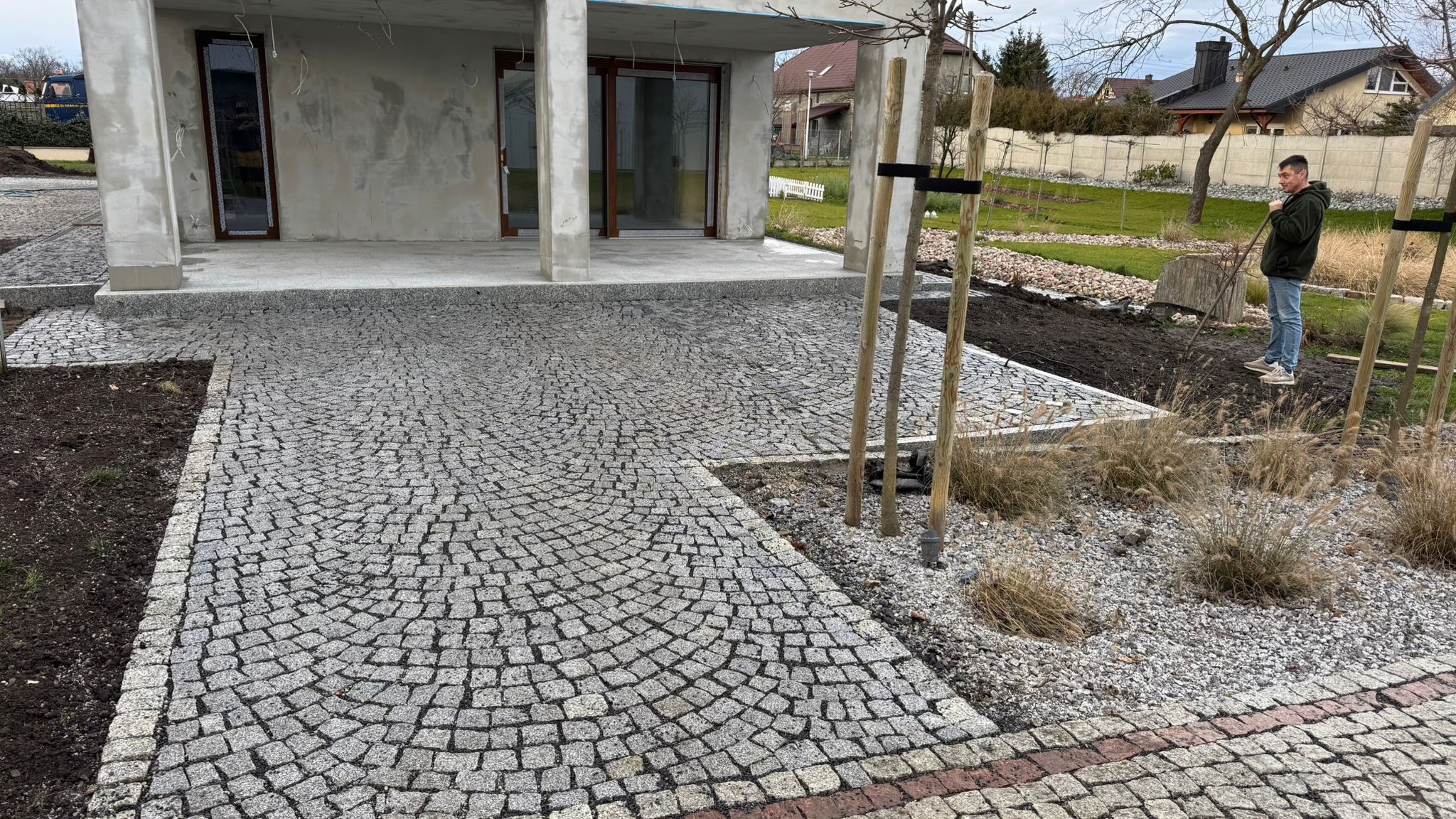 Szara kostka granitowa ułożona w łukowy wzór przed domem w trakcie prac wykończeniowych ogrodu, widoczny mężczyzna z grabiami.