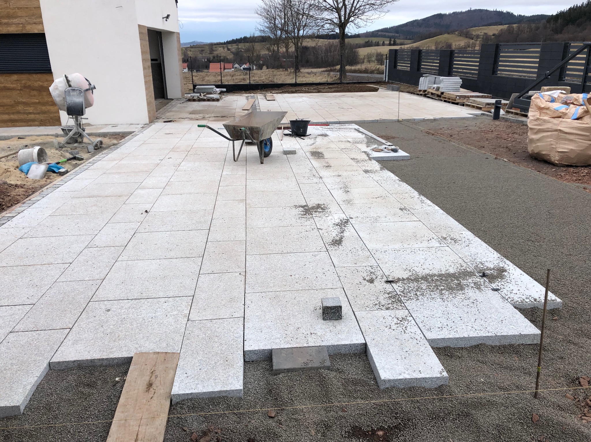 Trwa układanie jasnoszarej kostki granitowej na posesji, widoczna zaprawa, taczka, poziomica i mieszalnik do betonu w tle.