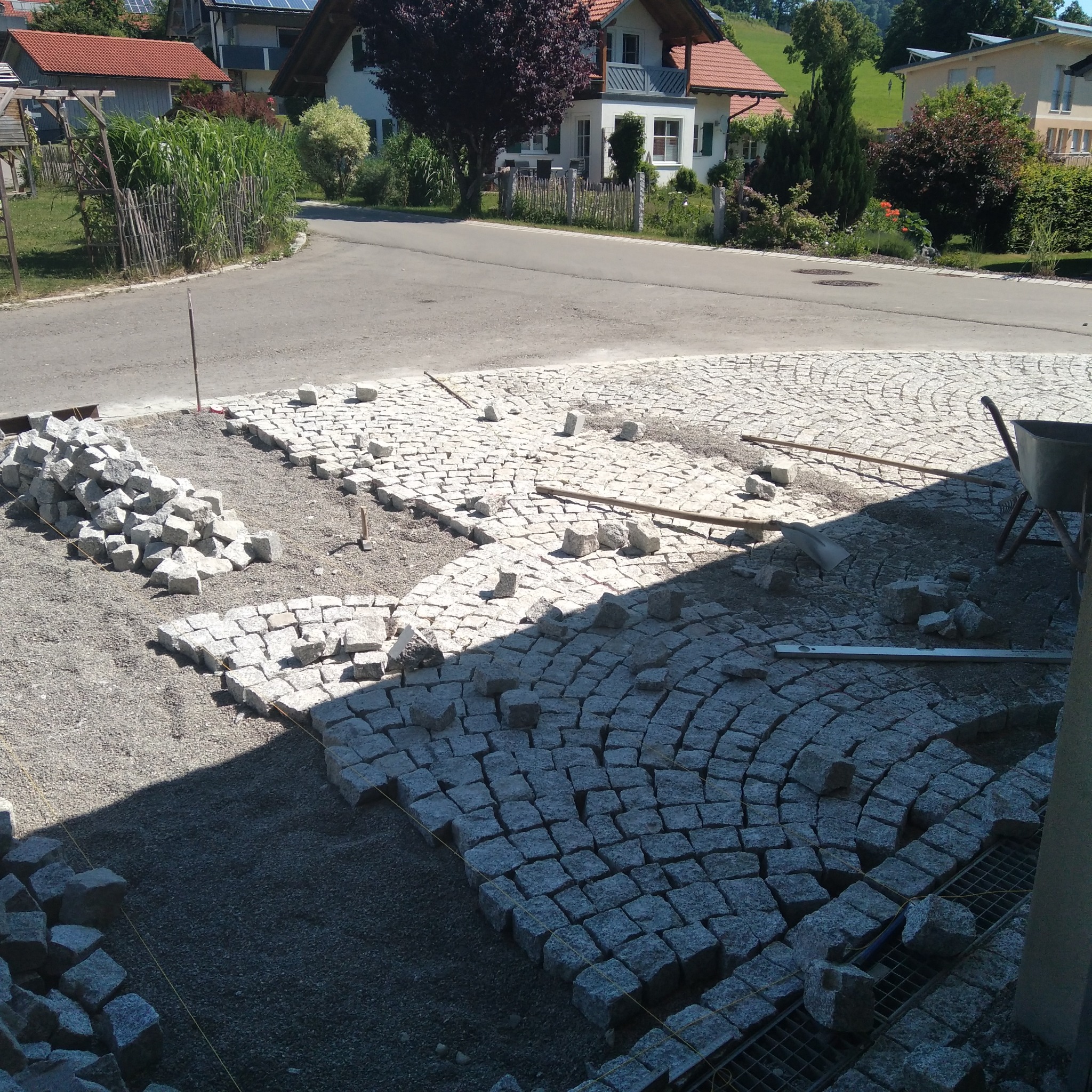 Układanie kostki granitowej w łuk, widoczny fragment ułożonej nawierzchni, stos kostki do ułożenia, narzędzia i taczka w tle, etap prac z widocznymi liniami pomocniczymi.