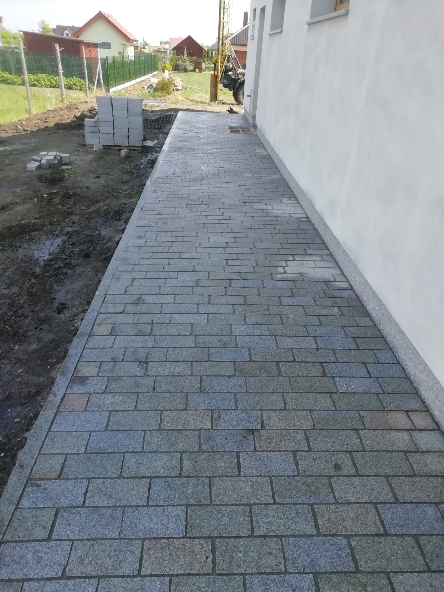 Ułożona ścieżka z szarej kostki granitowej prowadząca wzdłuż ściany budynku, z przygotowanym podłożem i ułożonymi kostkami w tle.